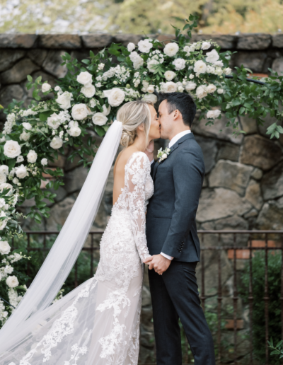 A bride and groom kiss in front of a stone wall adorned with white flowers. The bride wears a lace dress and veil, and the groom is in a suit.